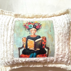 May include: Cream-colored decorative pillow with a printed image of a woman reading. The woman is seated in a chair, wearing a black dress and a floral crown. The pillow has a textured, fringed border.