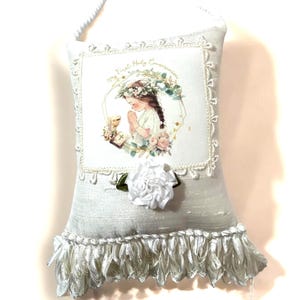 May include: A white pillow with a lace trim and a fringe border. The pillow features a printed image of a young girl in a white dress holding a chalice. The text "My First Holy Communion" is printed on the pillow.