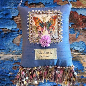 May include: A decorative blue pillow with a butterfly print, a lavender rose, and the text "The Best of Friends!". The pillow has a gold trim, a fringe at the bottom, and a hanging cord.