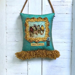 May include: Teal decorative pillow with a gold rope handle and fringe. The pillow features a framed image of birds perched on a branch. The frame is gold with a ruffled edge. The text "simple pleasures" is on the pillow.