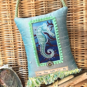 May include: A decorative pillow with a seahorse design. The pillow is light blue with a green trim and a framed image of a seahorse. The text "The Sea Calls To Me" is on the pillow. A green rope handle is attached.