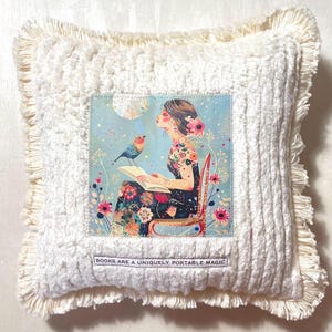 May include: Square decorative pillow with a cream-colored, textured cover and fringe trim. The center features a printed image of a woman reading with a bird, flowers, and the text "BOOKS ARE A UNIQUELY PORTABLE MAGIC."