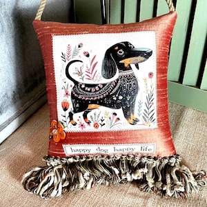 May include: Decorative pillow with a dachshund illustration. The pillow has a rust-colored border and a white center with a black and tan dachshund surrounded by flowers. The text "happy dog happy life" is at the bottom.
