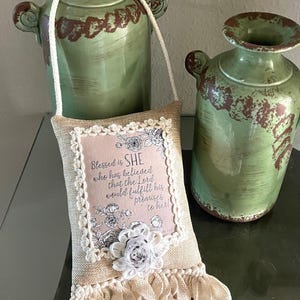 May include: A beige burlap pillow with a white lace trim and a white flower embellishment. The pillow has a pink and white floral print with the text "Blessed is SHE who has believed that the Lord would fulfill his promises to her!"