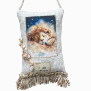 May include: A decorative pillow with a sleeping lion illustration. The pillow has a cream-colored woven texture, a cream-colored fringe, and a cream-colored rope handle. The illustration features a lion with a brown mane and the text "just dream".