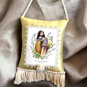 May include: A decorative, yellow silk pillow with a printed image of a religious figure holding a lamb, surrounded by flowers. The image is framed with a white lace border. The pillow has a cream-colored fringe and a white rope handle.