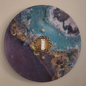 May include: A circular light switch plate featuring a marbled pattern in shades of purple, teal, and gold. The center has a white toggle switch surrounded by a metallic gold chain-link border, secured with two small gold-colored screws.