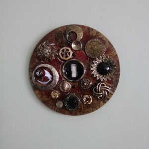 May include: A round, decorative wall art piece with a reddish-brown background. It features an array of vintage buttons and embellishments in various shapes, sizes, and metallic finishes, including gold, silver, and black. A central light switch is integrated into the design.