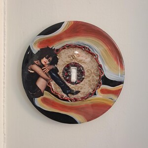 May include: A decorative light switch cover featuring a woman in a black dress and boots. The round cover has a black, red, and yellow swirl design. The switch is white.