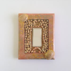 May include: Decorative light switch cover with a rectangular frame in shades of pink and gold. The frame is embellished with small, gold-colored stones and seashells. A white light switch is in the center, with gold accents.