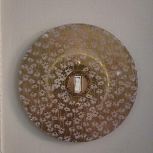 May include: A circular light switch cover plate featuring a gold background with a repeating white daisy floral pattern. The plate has a central toggle switch with a white lever and is secured by two small gold screws positioned vertically.