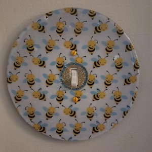 May include: A circular light switch plate featuring a repeating pattern of yellow and black cartoon bees on a light blue background. The center includes a white toggle switch surrounded by a silver glitter border and two yellow mounting screws.