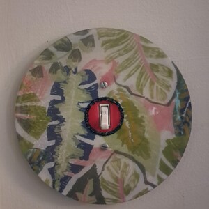 May include: A circular light switch plate featuring a botanical pattern of green, pink, and dark blue leaves. The plate has a central white toggle switch set within a red circular border, secured by two silver screws positioned above and below.