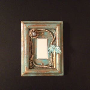 May include: Decorative light switch cover with a rectangular frame in distressed teal and brown. The frame is adorned with beaded accents, a palm tree, and a light switch. The cover is mounted on a black wall.