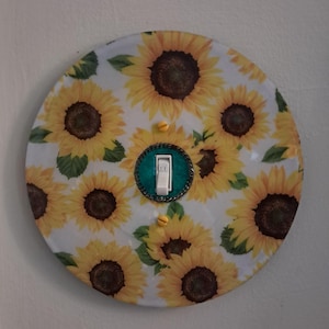 May include: A circular light switch cover plate featuring a repeating pattern of yellow sunflowers with dark brown centers on a white background. The center includes a white toggle switch surrounded by a teal border and two yellow screws.