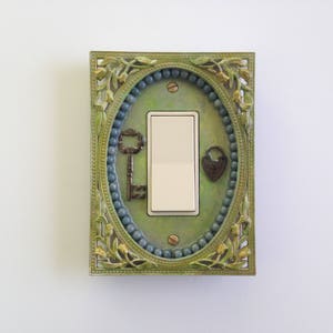 May include: An antique-style light switch cover with a decorative frame in shades of green and gold. The cover features an oval design with a beaded border, a key, and a heart-shaped lock. The switch itself is a standard rectangular shape.