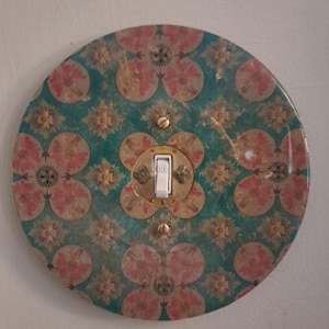 May include: A circular light switch plate featuring a repeating pattern of pink floral motifs and gold-toned starburst designs on a teal background. The plate is secured with two metal screws and has a central white toggle switch labeled OFF.