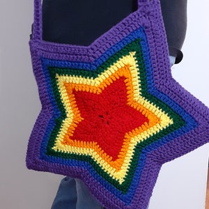 May include: A handmade, star-shaped crochet bag in a vibrant rainbow of colors. The bag features a purple border and strap, with a central star design in red, orange, yellow, green, and blue. The bag is worn over the shoulder.