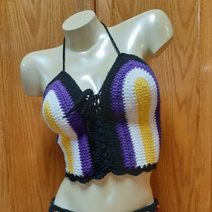 Pode incluir: Um top de halter de crochê com um padrão listrado roxo, branco e amarelo. O top tem um detalhe de renda preta na frente.