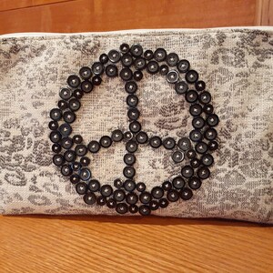 Puede incluir: Un embrague de tela blanca con cierre de cremallera y un diseño de signo de paz hecho de botones negros.