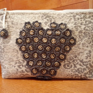 Può includere: Una pochette beige con cerniera e un design a forma di cuore realizzato con fiori all'uncinetto grigi e bottoni argentati.