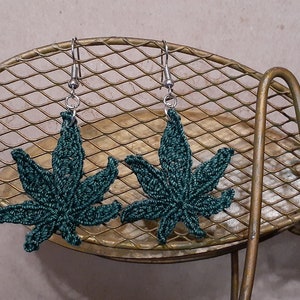 Op de afbeelding: Een paar groene gecrochete marihuana-bladeren oorbellen met zilveren haakjes. De oorbellen hangen aan een gouden metalen rek.