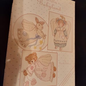 Peut inclure: Un livre de patrons de point de croix vintage intitulé "Priscilla's Babyland". La couverture présente trois motifs encadrés au point de croix : un lapin, un ange et un ours en peluche, tous dans des couleurs pastel douces. Le fond est rose clair avec de petits motifs de cœurs.