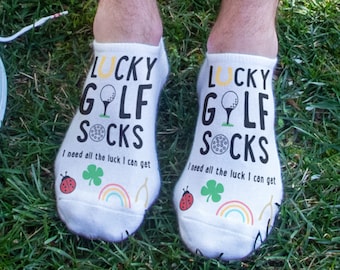 May include: A pair of white ankle socks with a golf ball and horseshoe design. The socks have the text "Lucky Golf Socks" and "I need all the luck I can get".
