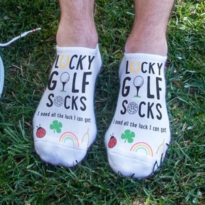 May include: A pair of white ankle socks with a golf ball and horseshoe design. The socks have the text "Lucky Golf Socks" and "I need all the luck I can get".