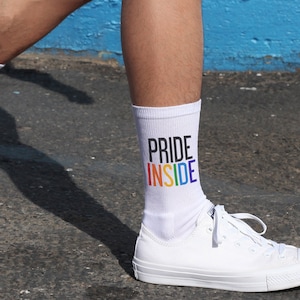 May include: White socks with the words "PRIDE INSIDE" in rainbow colors. The socks are being worn with white sneakers.