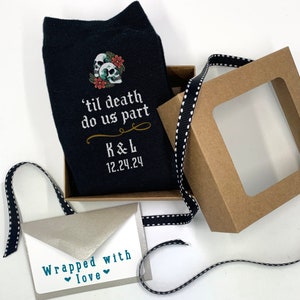 May include: A pair of black socks with a skull and floral design. The socks have the text "'til death do us part" and "K&L 12.24.24" printed on them. The socks are in a brown box with a clear window. A silver envelope with the text "Wrapped with love" is also in the image.