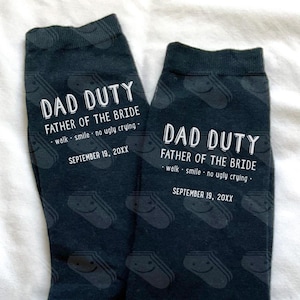 May include: A pair of black socks with a white pattern of smiling faces. The socks have the text "Dad Duty Father of the Bride" with the date "September 19, 20XX".