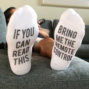 May include: A pair of white socks with black text. One sock says "If you can read this" and the other sock says "Bring me the remote control."