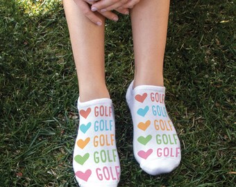 May include: White ankle socks with a colorful heart and "GOLF" design. The socks are worn on a person's feet.
