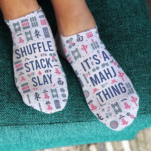 May include: White ankle socks with a Mahjong tile pattern and the phrases "SHUFFLE STACK SLAY" and "IT'S A MAHJ THING" in navy blue. The socks are decorated with red and pink accents.