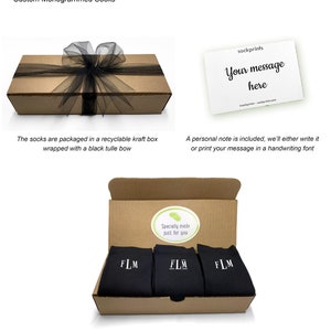 May include: A brown cardboard box with a black tulle bow, containing three pairs of black socks with white embroidered initials "FLM". The box is open and the socks are visible. The box has a sticker that says "Specially made just for you".