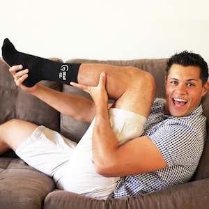 May include: A man wearing white shorts and a black and white checkered shirt is sitting on a brown couch. He is holding his leg up and pulling up a black sock with the text "GEAR CO." on it.