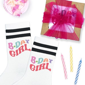 May include: A pair of white socks with black stripes and the words "B-Day Girl" in colorful letters. The socks are next to a small gift box with a pink ribbon and a cupcake with pink frosting.