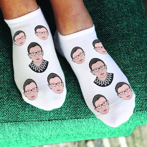 May include: A pair of white ankle socks with a repeated image of Ruth Bader Ginsburg. The socks feature a black and white illustration of Ginsburg wearing a black robe and a necklace with a symbol of female equality.