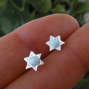 May include: A pair of star-shaped stud earrings. Each earring features a silver-colored, six-pointed star design with a light blue, oval-shaped stone in the center. The earrings are small and delicate, suitable for everyday wear.