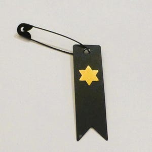 May include: A black tag with a gold Star of David design, attached to a black safety pin. The tag has a pointed bottom and a hole at the top for the pin. The background is a solid white.