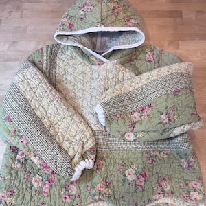 Peut inclure: Un pull à capuche matelassé en tissu vert sauge avec un motif floral rose et jaune, agrémenté de panneaux à motif pied-de-poule noir et blanc. Le vêtement présente des manches longues avec des poignets élastiques blancs et une capuche bordée de blanc.