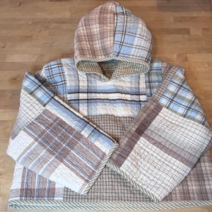 May include: A hooded pullover made from quilted fabric featuring a patchwork design of various plaid patterns in shades of light blue, white, brown, and tan. The garment has long, wide sleeves and a hood with a visible stitched grid texture throughout.