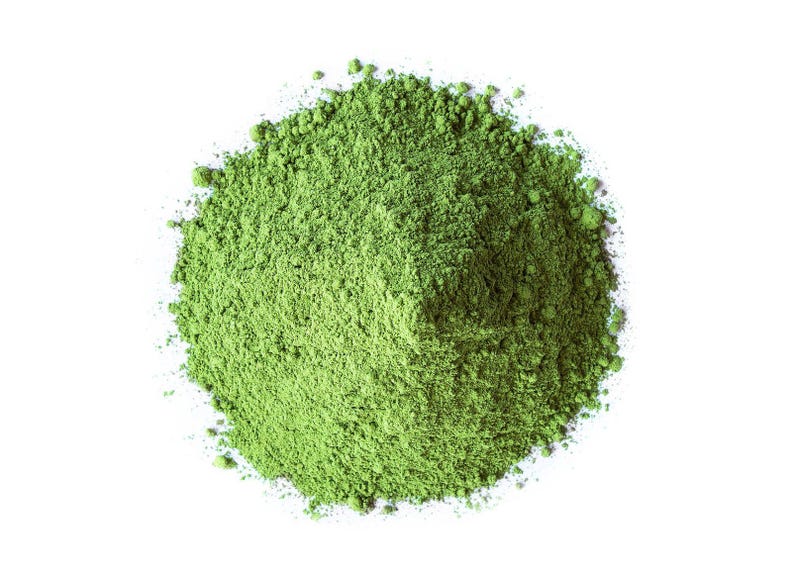 May include: A pile of finely ground, vibrant green powder against a stark white background. The powder appears to be matcha or a similar finely milled green tea, with a slightly textured appearance.