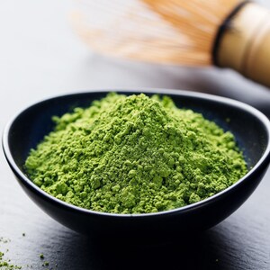 May include: A close-up shot of a black bowl filled with vibrant green matcha powder. A bamboo whisk rests in the background. The finely ground tea powder is a vivid green color, suggesting freshness and quality. The image evokes a sense of calm and wellness.