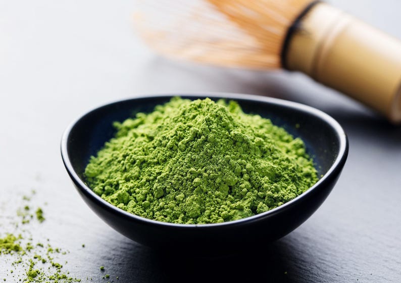 May include: A close-up shot of a black bowl filled with vibrant green matcha powder. A bamboo whisk rests in the background. The finely ground tea powder is a vivid green color, suggesting freshness and quality. The image evokes a sense of calm and wellness.