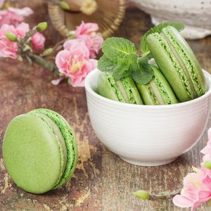 May include: A close-up of green macarons, some in a white bowl, others scattered on a wooden surface. Pink flowers and sprigs of mint add color. The macarons have a light green color and a textured surface.