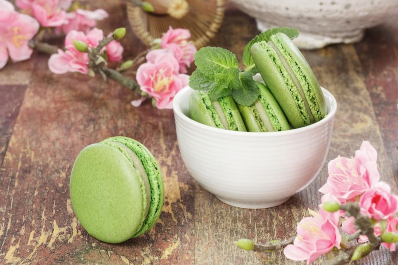 May include: A close-up of green macarons, some in a white bowl, others scattered on a wooden surface. Pink flowers and sprigs of mint add color. The macarons have a light green color and a textured surface.