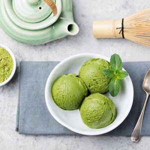 May include: Overhead shot of matcha-themed items. Three scoops of green ice cream in a white bowl with mint leaves, a small bowl of matcha powder, a green teapot, and a bamboo whisk. A silver spoon rests on a gray napkin.