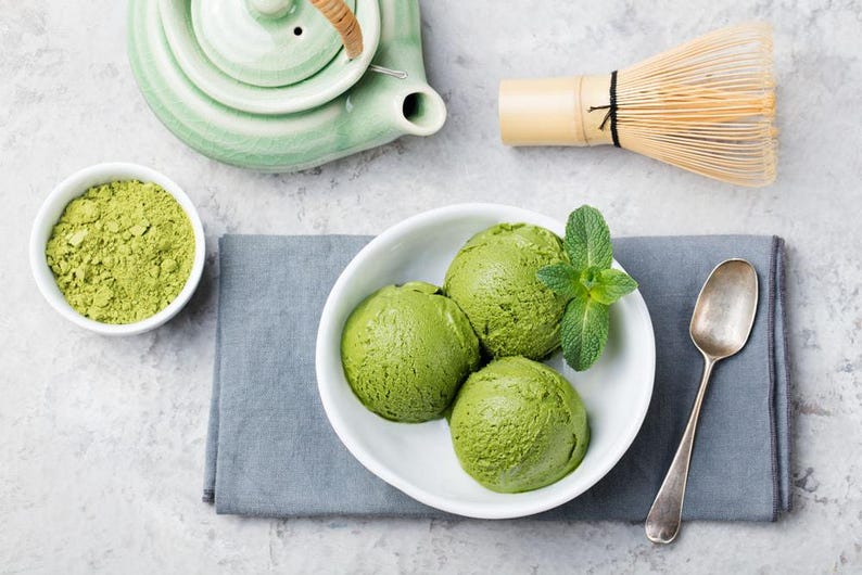May include: Overhead shot of matcha-themed items. Three scoops of green ice cream in a white bowl with mint leaves, a small bowl of matcha powder, a green teapot, and a bamboo whisk. A silver spoon rests on a gray napkin.